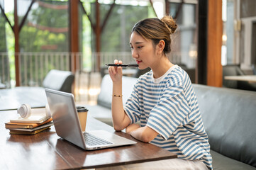 Asian young adult woman holding fountain pen and looking at laptop while sit at wooden table in cafe