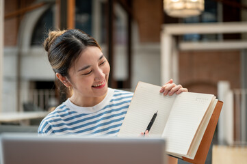 Fototapeta premium Smiling asian young woman holding pen pointing in notebook while sits on sofa across laptop in cafe.