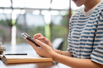 Close up of a woman holding and looking at smartphone over book while sitting at wooden table in...