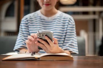 Close up of a woman holding and looking at phone over open book while sits at wooden table in a cafe