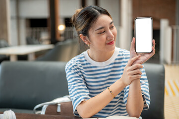 Asian young woman holding and showing white screen smartphone while sitting at wooden table in cafe.