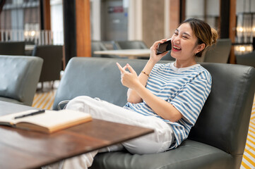 Asian young woman laughing during a phone call while sits on sofa with book on wooden table in cafe.