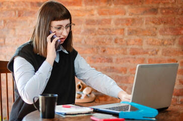 Young female employee working from home office with a laptop and talking on the phone looks...