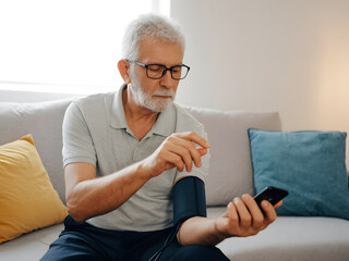 Senior Man Measuring Blood Pressure While Using Smartphone on a Comfortable Sofa. Elderly man seated on a sofa measuring his blood pressure while checking a smartphone, representing healthcare.