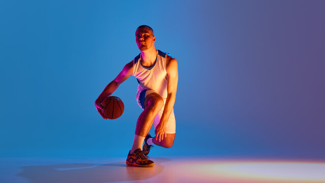 Basketball athlete dribbling ball against vibrant neon background. Concept of colored lighting, movement isolation, urban sports fashion, and stylized male athlete presence.
