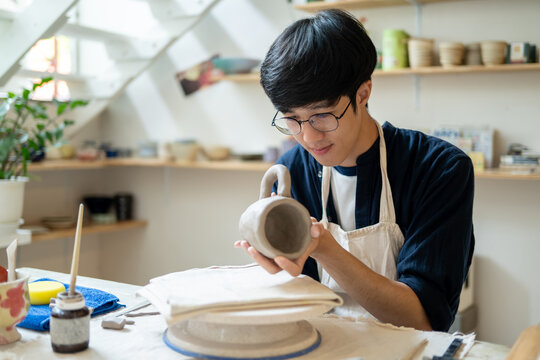 Young man potter artist holding and looking at clay mug at working table in pottery class or studio - Powered by Adobe