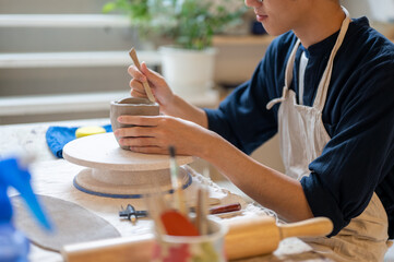Young pottery artist using a throwing tool to make clay mug at working table in a class or studio.