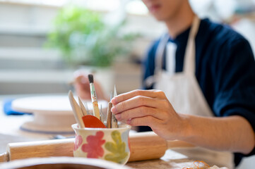Young pottery artist taking a painting brush from ceramic cup on working table in a class or studio.