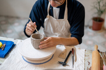 Glasses man pottery artist using a throwing tool on clay mug at working table in a class or studio.