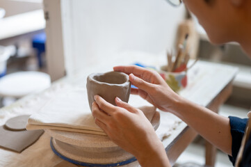 Close up of young pottery artist's hand molding on a clay mug at working table in a class or studio.