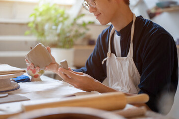 Young pottery artist holding and looking at clay mug with handle at working table in class or studio