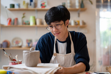 Young man pottery artist holding and looking at clay mug or handle at working table in class or studio.