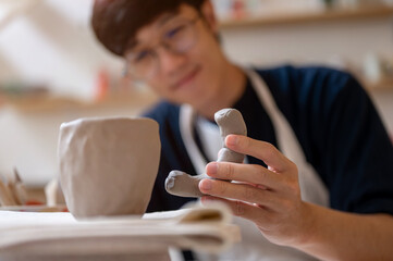 Young man pottery artist holding and looking at clay mug handle at working table in class or studio.