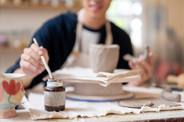 Potter artist holding brush over slip bottle for gluing clay mug at working table in class or studio
