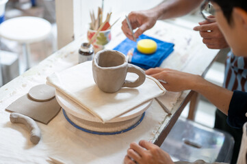 Pottery teacher and a student looking at clay mug over a throwing wheel on table in class or studio.