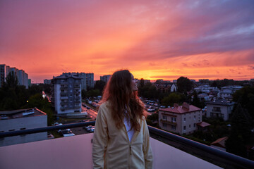 Young woman enjoying a vibrant sunset over an urban cityscape from a rooftop, with the sky...