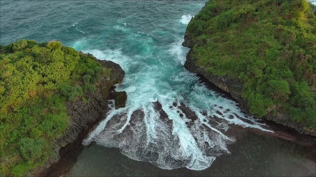 The aerial view shows waves continuously crashing into the gap between two cliffs covered with green trees in the afternoon.