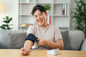 An Asian woman uses a portable blood pressure monitor in her living room. Health awareness concept.