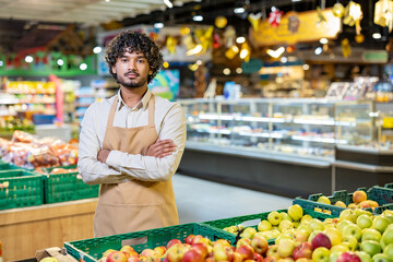 A supermarket employee stands confidently in front of a display of fresh produce with arms crossed, looking directly at the camera.