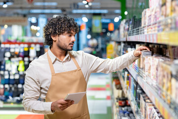 An employee in a beige apron checks products on a supermarket shelf while holding a tablet.
