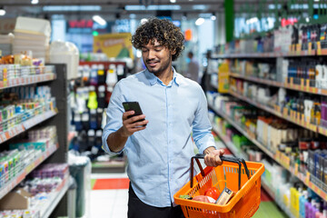 A man shops in a grocery store, using his phone while holding a basket with groceries. He's smiling.