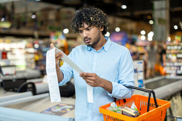 A man in a grocery store looks at a long receipt, seemingly surprised by the total cost of his purchase.