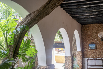 View of white arches framing a glimpse of outdoor seating, complemented by rough stone textures and vibrant green foliage, creating a tranquil scene, Lamu, Kenya.
