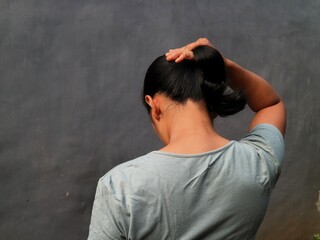 Asian Indonesian woman, seen from behind, is putting her hair up with a focused and casual gesture.
