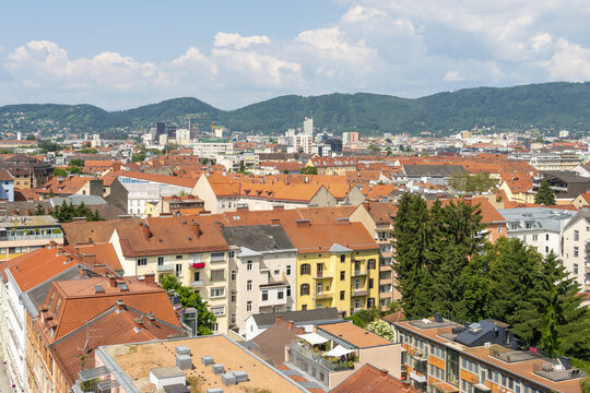 Fototapeta View of terracotta rooftops cascading under a bright sky, nestled against lush green hills and the city skyline, blending nature and urban life, Graz, Austria.