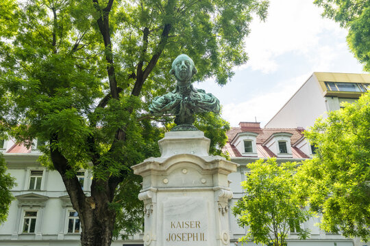 View of Kaiser Joseph II's bust stands proudly in a park, framed by lush green trees and elegant buildings under a bright sky, Graz, Austria.