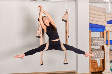 Woman performs aerial yoga in sunlit studio. Neutral-toned walls, wooden floor, vibrant fabric...