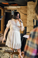Obraz premium Young adult woman with black hair passionately sings in rustic music studio. Warm lighting enhances cozy atmosphere, with guitarist in foreground adding dynamic energy