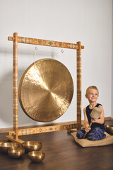 Young child seated on cushion, holding mallet beside large golden gong and arranged singing bowls....