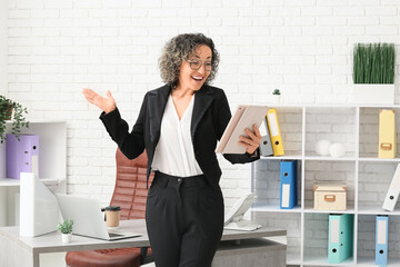 Surprised mature businesswoman in eyeglasses with tablet near workplace in modern office