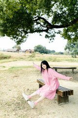 Naklejka premium Young Asian woman in pink dress sitting on a bench outdoors.