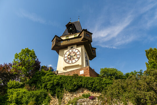View of the clock tower standing prominently, ivy crawling over the stone wall, contrasting against the vibrant blue sky, Graz, Styria, Austria.