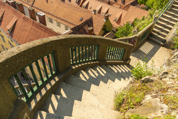 View of a winding stone staircase descends past a weathered concrete railing with green bars, offering glimpses of terracotta rooftops, Graz, Styria, Austria.