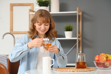 Young woman adding sweet honey into cup of tea in kitchen