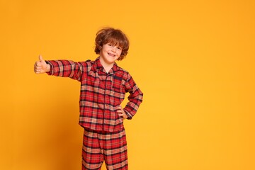 Cute little boy in pajamas showing thumbs up on yellow background. Space for text