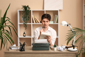 Young writer with notebook and vintage typewriter sitting at table in room