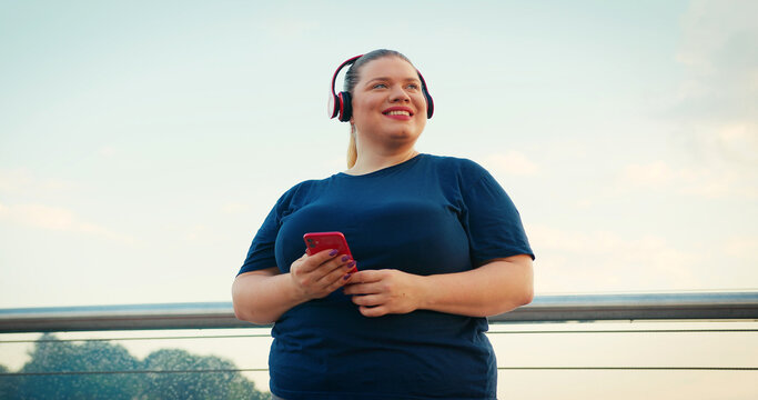Outdoor Portrait of Body Positive Overweight Woman Using Red Smartphone Phone Outdoors at City in the Morning. Wearing Red Headphones