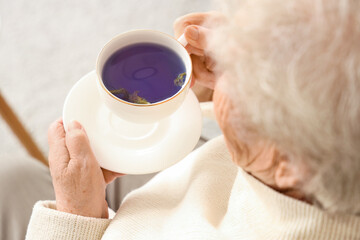 Senior woman with cup of butterfly pea flower tea at home, closeup