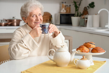 Senior woman with glass cup of butterfly pea flower tea and sweet pastry at table in kitchen