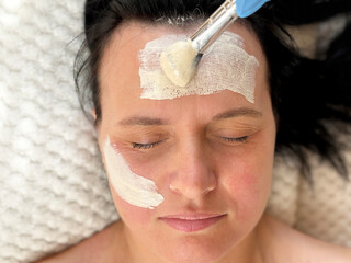 Mature woman with wrinkles lying on a white cosmetology table during a facial procedure. White clay...