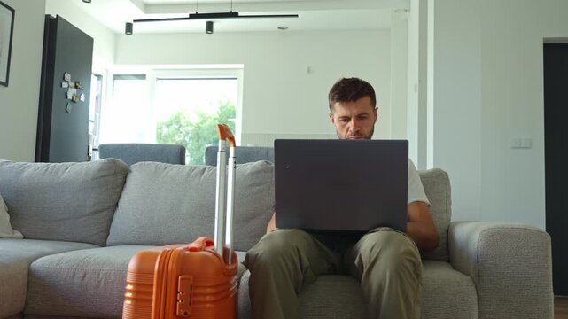 Adult man sitting on sofa in modern living room near orange suitcase and working on laptop. Concept of travel, remote work and digital nomad lifestyle