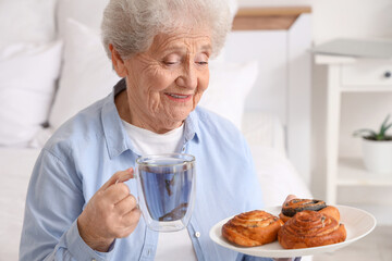 Senior woman with glass cup of butterfly pea flower tea and plate of sweet pastry in bedroom
