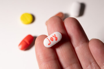 A hand holds a white pill inscribed with the word joy, surrounded by colorful pills. This...