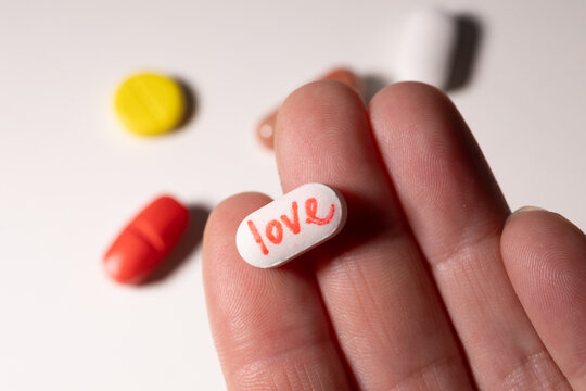 A hand holds a white pill marked with the word love, surrounded by vibrant capsules on a surface. The image symbolizes the complex relationship between addiction, joy, and wellness.