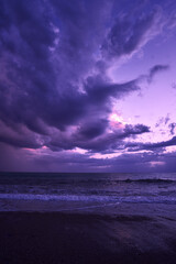 Vibrant seashore at dusk showcasing purple hues in the sky with dramatic clouds, reflecting on calm ocean waves, creating peaceful natural scene suitable for relaxation and contemplative themes.