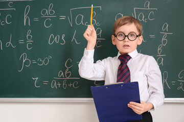 Cute schoolboy with clipboard having idea near chalkboard in classroom. Children's Inventor's Day
